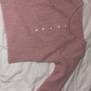 Rosa stickad tröja med pärldetaljer - Säljer en söt rosa stickad tröja med dekorativa pärlknappar framtill. Tröjan har en ribbad struktur och långa ärmar, perfekt för kyligare dagar. Den är både stilren och bekväm, ett måste i garderoben! 💖