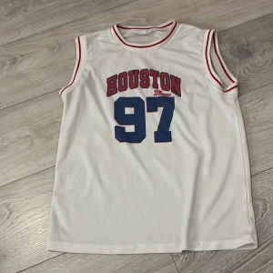 Vit ärmlös topp från Shein med Houston-tryck - Säljer en vit ärmlös topp från Shein med rött och blått Houston-tryck och siffran 97 på framsidan. Toppen har röda detaljer vid ärmhål och halsringning. Perfekt för en sportig look!