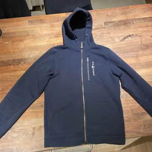 Mörkblå hoodie från Sail Racing - Säljer en snygg mörkblå hoodie från Sail Racing med dragkedja och broderad logga på bröstet. Perfekt för kyliga dagar med sin mjuka insida. Den är lite liten i sin storlek. Använd fåtal gånger och är i bra skick