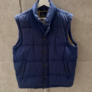 Blå quiltad väst från Fred Perry - Säljer en snygg blå quiltad väst från Fred Perry. Västen har en hög krage och knappar framtill. Den är perfekt för kyligare dagar och har en stilren design med diskret logga på bröstet.