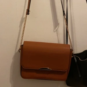 Brun axelväska med kedjedetalj - Snygg brun axelväska med stilren design och kedjedetalj på axelremmen. Perfekt för att ge en touch av elegans till din outfit. Väskan har en praktisk storlek för att bära dina nödvändigheter.