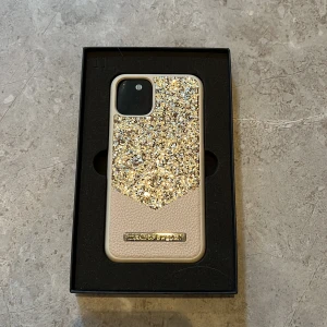 Glimrande mobilskal från iDeal of Sweden - Snyggt mobilskal från iDeal of Sweden för iPhone 11 Pro/XS/X. Skalet har en glittrig design i guld och beige med en elegant metallplatta. Perfekt för att ge din telefon en lyxig touch.