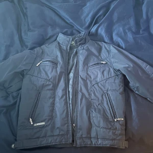 Svart jacka från North Sails - Säljer en svart jacka från North Sails med dragkedja och flera praktiska fickor. Jackan har långa ärmar och en stilren design, perfekt för kyligare dagar. Den är tillverkad i ett syntetmaterial som ger en lätt och bekväm känsla.