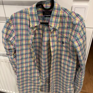 Rutig skjorta från Ralph Lauren - Snygg rutig skjorta från Ralph Lauren i en mix av färger som blå, gul, grön och rosa. Skjortan har en slim passform och är långärmad med knappar framtill. Perfekt för en stilren look. Pris går att diskutera då jag gärna vill bli av med den!
