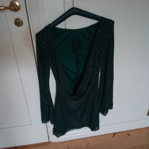 Glittrig grön klänning - Snygg grön klänning med glittriga detaljer och djup v-ringad rygg. Perfekt för en kväll ute! Klänningen har långa ärmar och en figurnära passform.