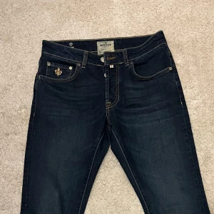 Mörkblå jeans från Morris - 29 32 Snygga mörkblå jeans från Morris med broderad detalj på fickan och klassisk femficksdesign. De har en midwaist passform och är perfekta för en stilren look. Bältet medföljer och ger en extra touch till outfiten.