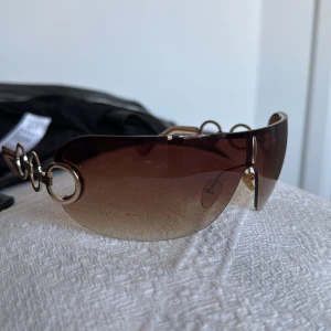 90s bruna solglasögon med gulddetaljer - Snygga solglasögon (riktiga från 90-talet) med bruna tonade linser och guldfärgade detaljer. Bågarna har en unik design med cirkulära länkar på sidorna som ger en elegant touch. Perfekta för en solig dag!