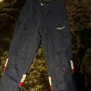 Blå mjukisbyxor från Adidas - Snygga blå mjukisbyxor från Adidas med vita och röda detaljer vid benen. Byxorna har elastisk midja och dragkedjefickor framtill. Perfekta för en avslappnad stil.