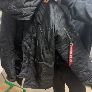 Svart parkajacka med pälsfoder  - Säljer en svart parkajacka från Alpha Industries med pälsfoder i huvan och orange insida. Jackan har flera fickor med dragkedja och knappar, samt en ikonisk röd tagg på ärmen. Perfekt för kalla dagar!