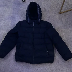 Svart pufferjacka från GANT - Säljer en snygg svart pufferjacka från GANT med huva och dragkedja. Jackan har märkets logga på ärmen och längs dragkedjan. Perfekt för kyligare dagar.