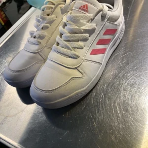 Vita sneakers från Adidas med rosa detaljer - Snygga vita sneakers från Adidas med rosa ränder och logga. Skorna har snörning och en klassisk design som passar perfekt till vardags. De är tillverkade i syntetmaterial för en hållbar och bekväm känsla.