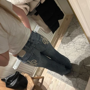 Blå jeans med broderad detalj - Snygga blå jeans med unika broderade detaljer på bakfickorna. De har en låg midja och en bootcut-stil som ger en avslappnad look. Perfekta för dig som vill ha något annorlunda i garderoben!