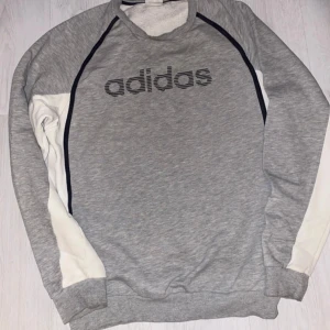 Grå sweatshirt från Adidas - Säljer en grå sweatshirt från Adidas med svart logga på bröstet. Tröjan har långa ärmar med vita och svarta detaljer. Perfekt för en avslappnad stil.