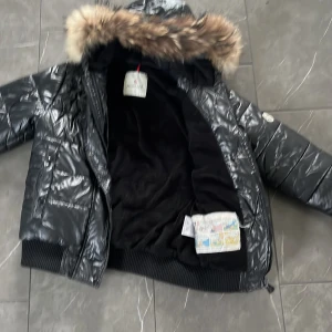 Svart pufferjacka från Moncler - Snygg svart pufferjacka från Moncler med pälsfodrad huva. Jackan har en glansig yta och dragkedja framtill. Perfekt för kyliga dagar.