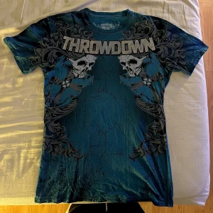 Blå t-shirt med tryck från Throwdown - Säljer en blå t-shirt från Throwdown med ett coolt tryck av dödskallar och texten 'THROWDOWN' på framsidan. T-shirten har korta ärmar och är perfekt för en avslappnad stil. Materialet är mjukt och bekvämt.