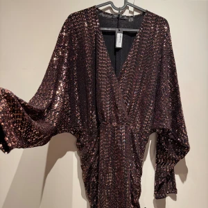 Svart glittrig klänning med paljetter i strl L - oanvänd! - Snygg svart klänning med glittrande paljetter och v-ringad design. Perfekt för en kväll ute med sina långa ärmar och figursydda passform. Klänningen har en elegant omlottstil som ger en sofistikerad look.