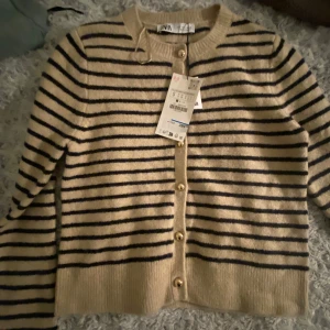 Beige och svart randig kofta från Zara - Säljer en snygg beige kofta från Zara med svarta ränder. Koftan har långa ärmar och knäpps med stiliga knappar framtill. Perfekt för en stilren look.