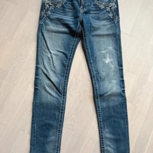 Blå jeans med broderade detaljer - Snygga blå jeans från Miss Me med unika broderade detaljer på bakfickorna. Jeansen har en skinny passform och är dekorerade med knappar i metall. Perfekta för dig som vill ha något extra i garderoben!