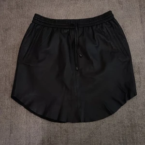 Svart skinn kjol från Second Female - Snygg svart skinn kjol från Second Female i en klassisk design. Kjolen har en elastisk midja med knytband och två sidofickor.