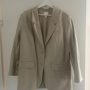 Beige kavaj från H&M - Säljer en stilren beige/grön kavaj från H&M i storlek L. Kavajen har en klassisk design med två knappar framtill och långa ärmar. Perfekt för en chic och avslappnad look. 🕶️