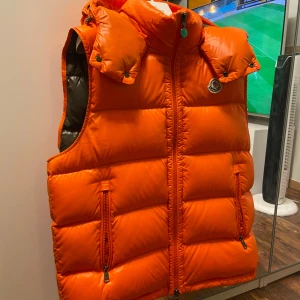 Moncler väst  - Säljer en snygg orange dunväst från Moncler med dragkedja och huva. Västen har två fickor med dragkedja framtill och Moncler-logga på bröstet. Perfekt för att hålla värmen med stil! Storlek 3 vilket motsvarar M/L. Kvitto från NK där den är inköpt finns och tillkommer, nypris 11000kr
