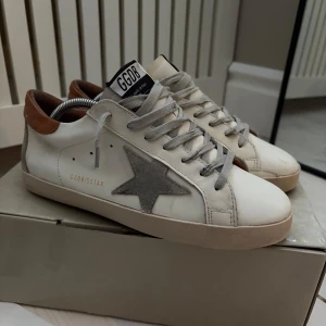 Golden Goose sneakers skor - Snygga Golden Goose sneakers i vitt med grå stjärndetalj på sidan. Skorna har bruna detaljer vid hälen och grå snörning. Perfekta för en stilren look med en touch av unik design. Alla tillbehör följer med. Super bra skick! Pris kan diskuteras
