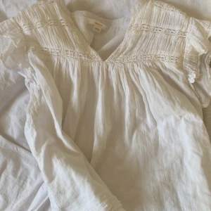 Vit blus från Gina Tricot - Säljer en söt vit blus från Gina Tricot med v-ringning och spetsdetaljer. Blusen har korta ärmar och en luftig passform, perfekt för varma dagar. Den är gjord i ett lätt och bekvämt material.