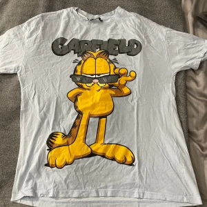 Vit Garfield t-shirt från Mister - Säljer en vit t-shirt med Garfield-tryck från Mister. T-shirten har en stor bild av Garfield med solglasögon på framsidan. Den är kortärmad och tillverkad i bomull, perfekt för en avslappnad stil.