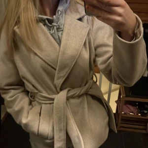 Beige kappa från Vero Moda i Storleken S - Säljer en stilren beige kappa från Vero Moda. Den har ett omlott-design med ett knytbälte i midjan. Perfekt för att hålla sig varm och snygg under kyligare dagar. Passar bra till både vardag och mer uppklädda tillfällen.💓💓