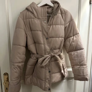 Beige quiltad pufferjacka med bälte - Säljer en superfin beige pufferjacka som är perfekt för kyliga dagar. Jackan är quiltad och har ett snyggt bälte i midjan för en mer definierad passform. Den har en huva och stängs med både dragkedja och knappar. Perfekt för höst och vinter!