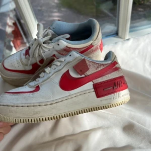 Nike Air Force 1 med röd detalj - Säljer ett par Nike Air Force 1 sneakers i vitt med röda detaljer. Skorna har en klassisk design med snörning och en robust sula. De är lite smutsiga men i bra skick. Perfekta för både vardag och fest!