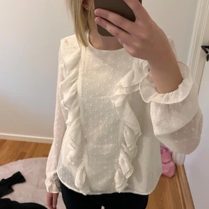 Vit blus med volangdetaljer - Säljer en söt vit blus med volangdetaljer. Blusen har långa ärmar och ett diskret mönster i tyget. Perfekt för både vardag och festliga tillfällen! En liten fläck men inget som syns tydligt. Köpt från Gina Young storlek 158.