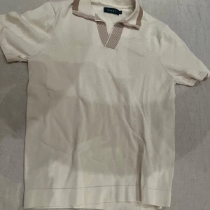 Beige pikétröja från Riley - Säljer en stilren beige pikétröja från Riley med korta ärmar och en klassisk krage i brunt. Perfekt för en avslappnad men ändå snygg look. Tröjan har en mjuk och bekväm känsla.