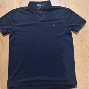 Mörkblå pikétröja från Ralph Lauren - Snygg mörkblå pikétröja från Ralph Lauren med klassisk krage och knappdetalj. Den har en broderad grön logotyp på bröstet och är kortärmad. Perfekt för en stilren look. Slim fit och riktigt skön inför sommaren. Priset är ej huggit i sten. Skriv om ni har några funderingar!
