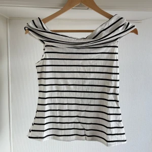 Randig offshoulder topp M oanvänd  - Snygg vit och svart randig offshoulder topp med en elegant krage. Perfekt för en stilren look. Toppen har en bekväm passform och är tillverkad i ett mjukt material.