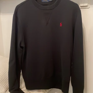 Svart tröja från Ralph Lauren - Snygg svart tröja från Ralph Lauren med det klassiska röda logotypen på bröstet. Tröjan har långa ärmar och en rund halsringning. Perfekt för en stilren look. Snabb affär= parfym sample