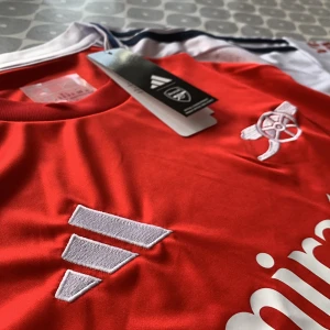 Röd Arsenal fotbollströja från Adidas - Snygg röd Arsenal fotbollströja från Adidas med vit och blå detaljering. Tröjan har korta ärmar och klubbens emblem broderat på bröstet. Perfekt för alla Arsenal-fans som vill visa sitt stöd. Officiellt licensierad produkt med 'Emirates Fly Better' tryckt på framsidan.