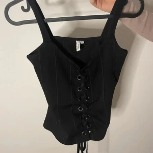 Svart korsett med snörning - Snygg svart korsett med snörning framtill. Perfekt för en kväll ute eller att styla med en kavaj. Den har breda axelband och en figurnära passform. Perfekt fest topp från NELLY