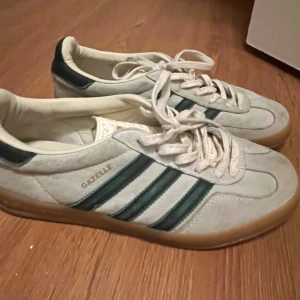 Adidas Gazelle sneakers - Säljer ett par klassiska Adidas Gazelle sneakers i vitt med mörkgröna ränder. Skorna har en snygg retrodesign med snörning och en gummisula som ger bra grepp. Perfekta för en avslappnad stil.