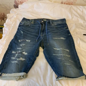 Blå jeansshorts med slitningar - Säljer ett par coola blå jeansshorts med slitningar och snörning i midjan. Shortsen har en avslappnad stil med upprullade benslut och klassiska femficksdetaljer. Perfekta för en casual look! 🕶️