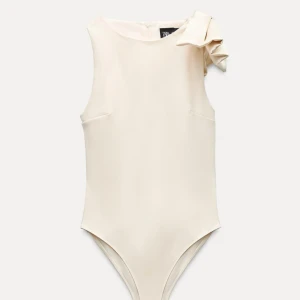 Body med rosett - Snygg beige bodysuit från Zara med en elegant rosett på axeln. Den är ärmlös och har en stilren design som passar perfekt för olika tillfällen. Perfekt för dig som vill ha en chic och modern look.