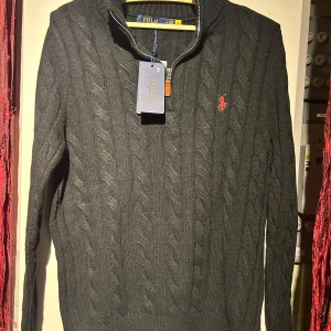 Grå stickad tröja från Polo Ralph Lauren - Snygg grå stickad tröja från Polo Ralph Lauren med dragkedja och kabelstickat mönster. Den har en liten röd logga på bröstet och en hög krage. Perfekt för kyliga dagar! OBS! Priset är inte hugget i sten. Hör av er vid minsta fundering 🍾