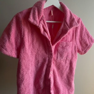 Rosa frottéset från NLYbeach - Säljer en häftigt rosa set i frotté från NLYbeach. Tröjan har korta ärmar och knappar framtill samt en praktisk bröstficka. Tillhörande shorts med fickor. Perfekt för en avslappnad stil och väldigt skönt att ha på sig. Aldrig använd, därav snöret i tröjan från prislappen.