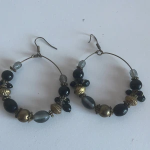 Örhängen med pärlor i svart och guld - Snygga örhängen med en mix av svarta och guldiga pärlor. De har en rund form och hängs i örat med en krok. Perfekta för att ge en elegant touch till din outfit.