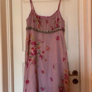 Rosa blommig klänning från KappAhl - Säljer en söt rosa klänning från KappAhl med blommigt mönster och silvriga detaljer. Klänningen har smala justerbara axelband och en dragkedja på sidan. Perfekt för sommarens festligheter! 🌸✨