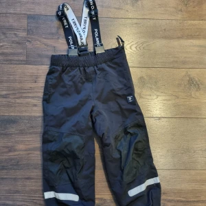 Svarta skalbyxor från Polarn O. Pyret. Mycket fina - Säljer ett par svarta skalbyxor / täckbyxor från Polarn O. Pyret med justerbara och avtagbara hängslen, reflexdetaljer, dragkedja på ena sidan i midjan för extra enkelt av och påtagande. Justerbar midja och elastiska benslut. Perfekta för utelek. Mycket fina. Strax dags byta fothällor.