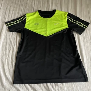 Svart och neongul t-shirt från Nike - Säljer en cool svart och neongul t-shirt från Nike. Den har korta ärmar och är tillverkad i 100% polyester. Perfekt för träning eller en sportig look. Nike-loggan syns både på bröstet och ärmarna.