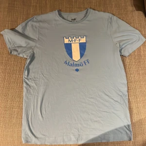 Ljusblå t-shirt med Malmö FF-logga - Säljer en ljusblå t-shirt med Malmö FF:s ikoniska logga tryckt på framsidan. Perfekt för alla fotbollsfans som vill visa sitt stöd för laget. T-shirten har en klassisk passform och korta ärmar.