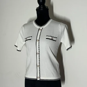 Vit kortärmad blus med svarta detaljer - Elegant vit blus med korta ärmar och svarta kantdetaljer. Den har dekorativa knappar framtill och två fickor med knappdetaljer. Perfekt för en stilren look.