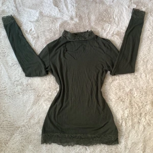 Mörkgrön ribbad topp med spetsdetaljer - Säljer en elegant mörkgrön ribbad topp från Gina Tricot. Toppen har långa ärmar och vacker spetsdetalj vid halsen och ärmsluten. Perfekt för en stilren look.